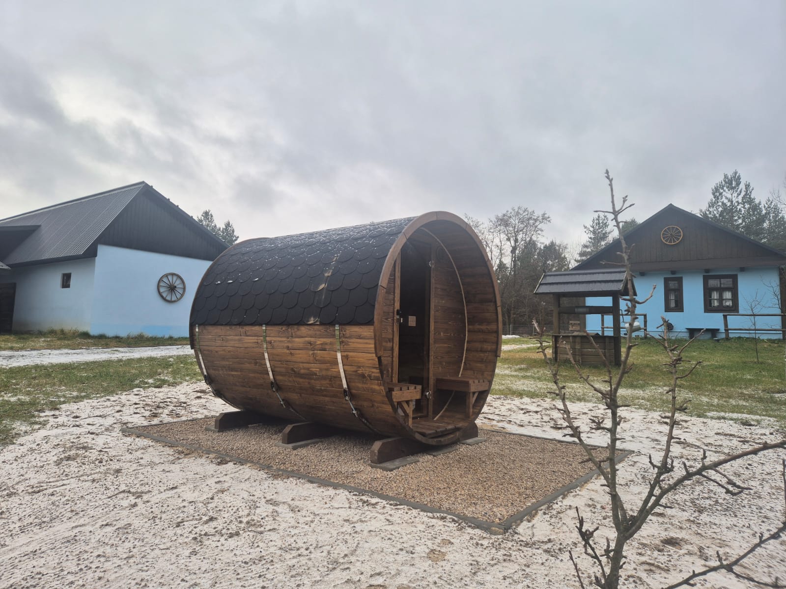 Drewniana sauna beczkowa na zewnątrz, otoczona żwirową powierzchnią i trawiastym terenem, z domami w tle i pochmurnym niebem.