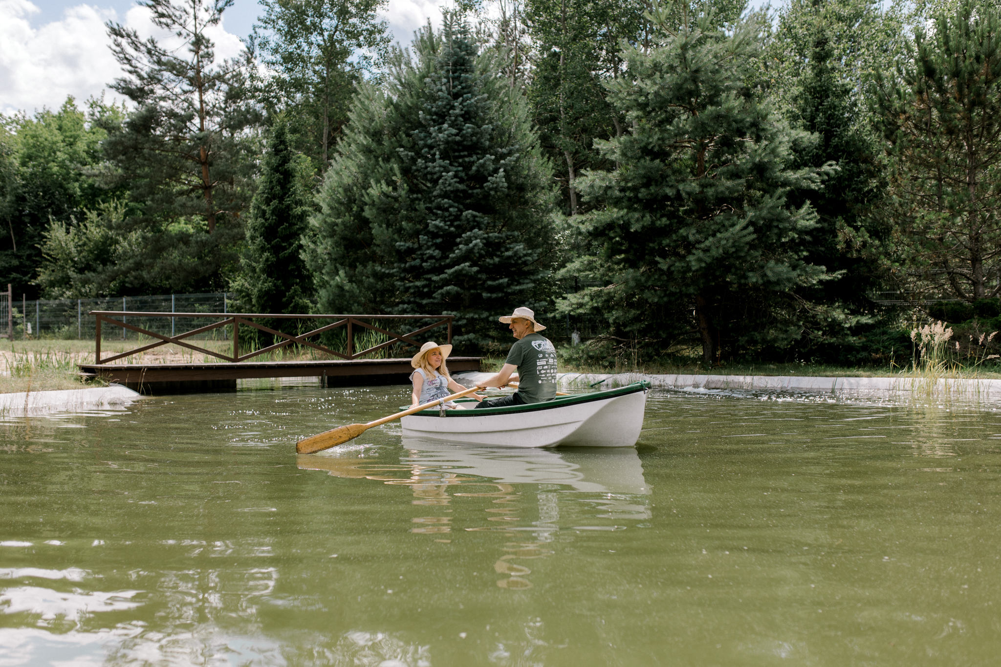 Dwie osoby wiosłujące w małej łodzi na jeziorze, otoczone zielonym lasem i drewnianym mostem, pod jasnym niebem z chmurami.