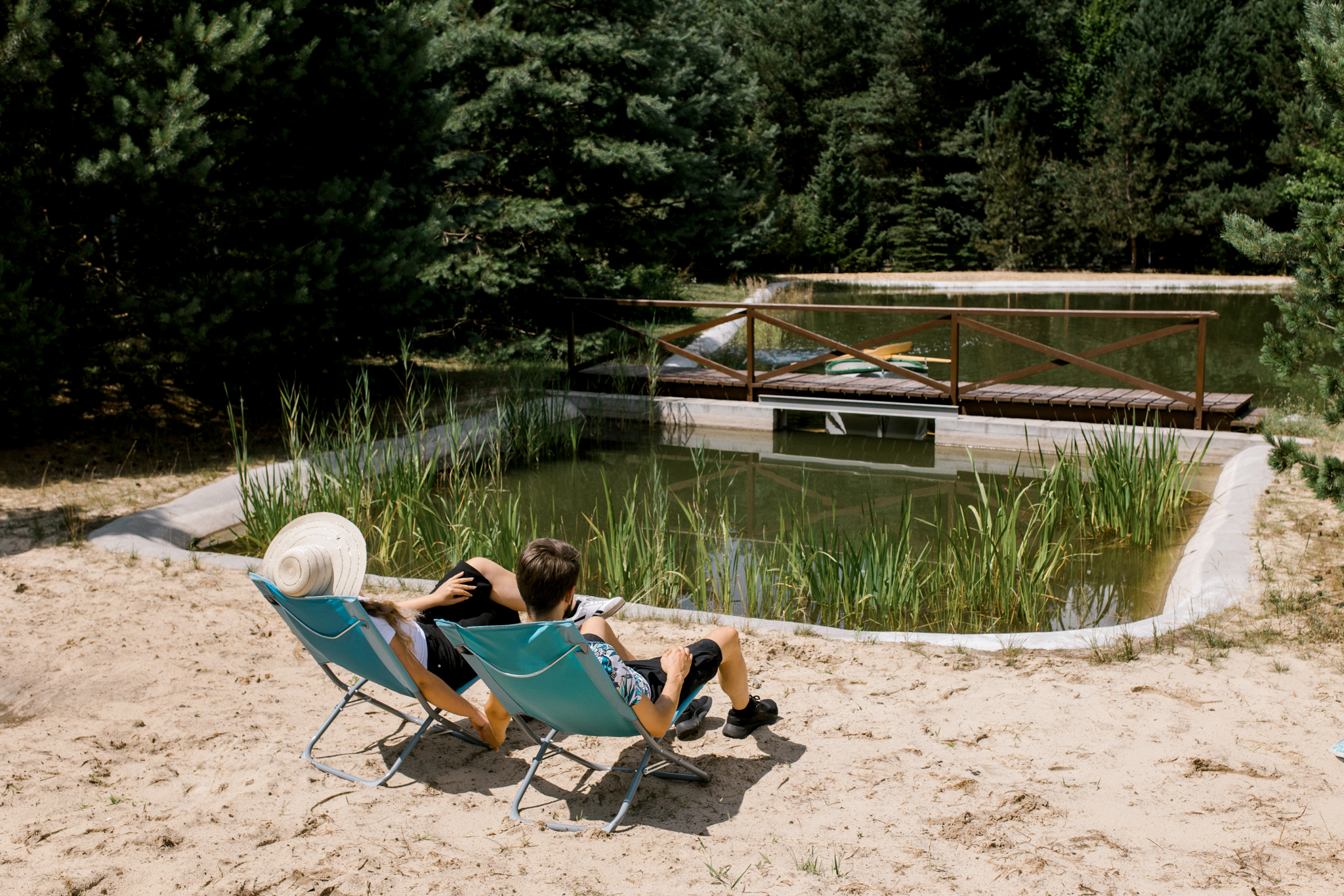 Dwie osoby odpoczywające na niebieskich krzesłach nad brzegiem jeziora z drewnianym mostem i lasem w tle.