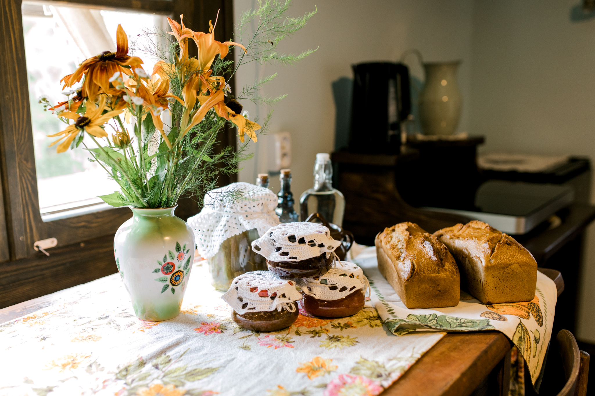Stół z kolorowym obrusem, domowym chlebem, słoikami dżemu i bukietem żółtych kwiatów w wazonie przy oknie.