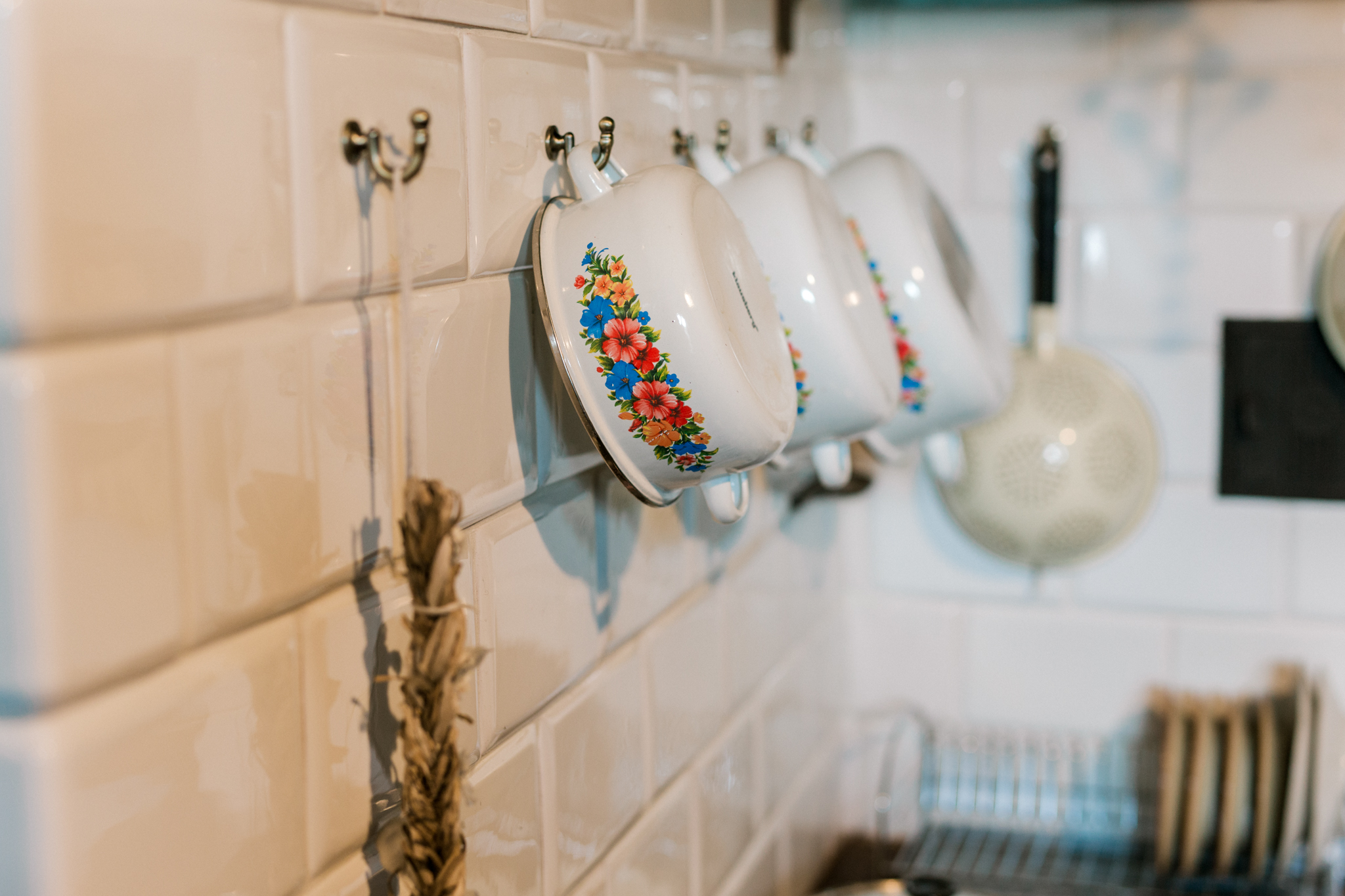 Kolorowe kubki emaliowane wiszące na białych kafelkach w tradycyjnej kuchni, z suszonymi ziołami dla dekoracji wnętrzarskiej.