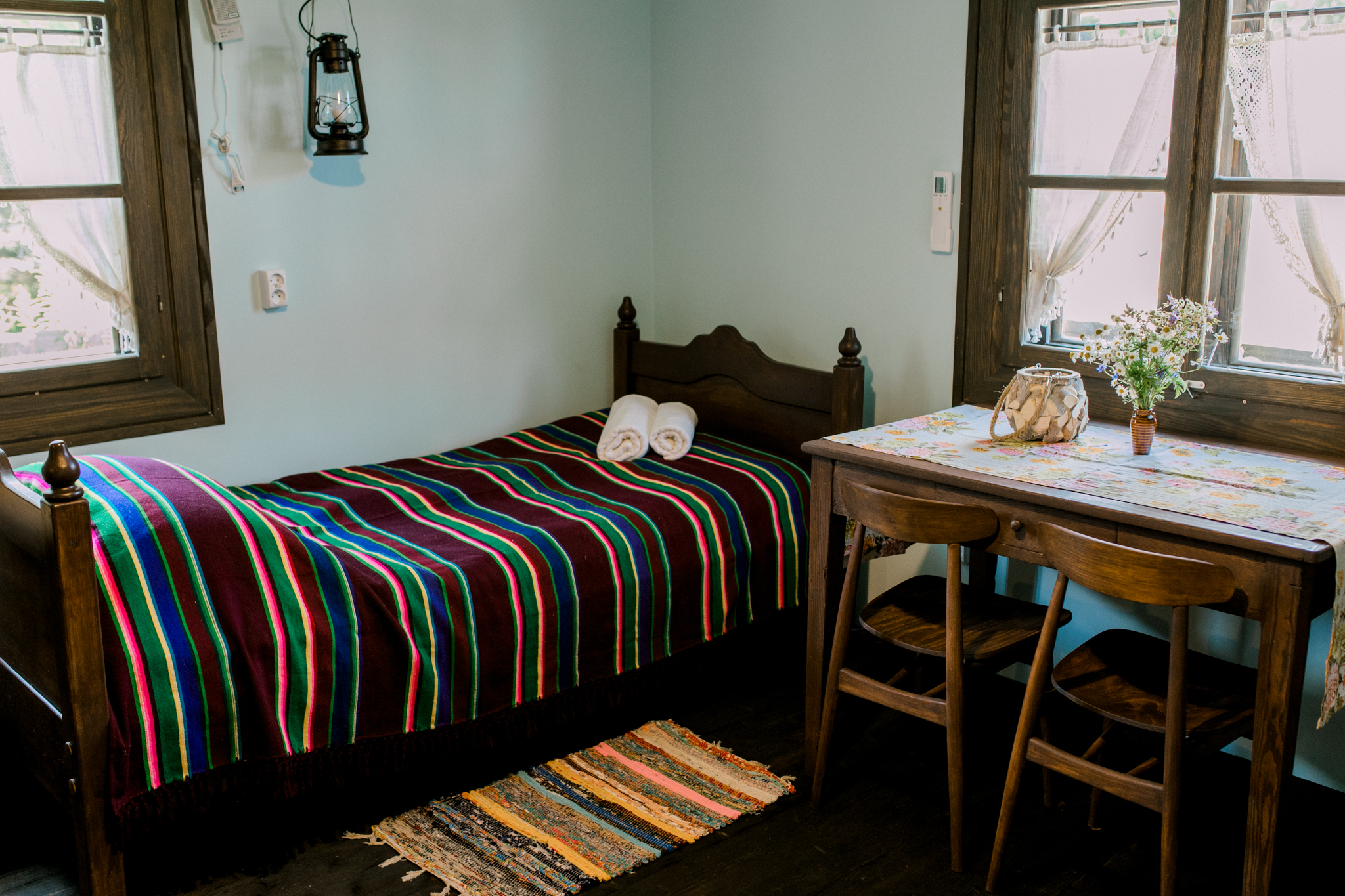 Przytulny pokój sypialny z kolorowym kocem na łóżku, ręcznikami i małym stołem z kwiatami w stylu folkloru.