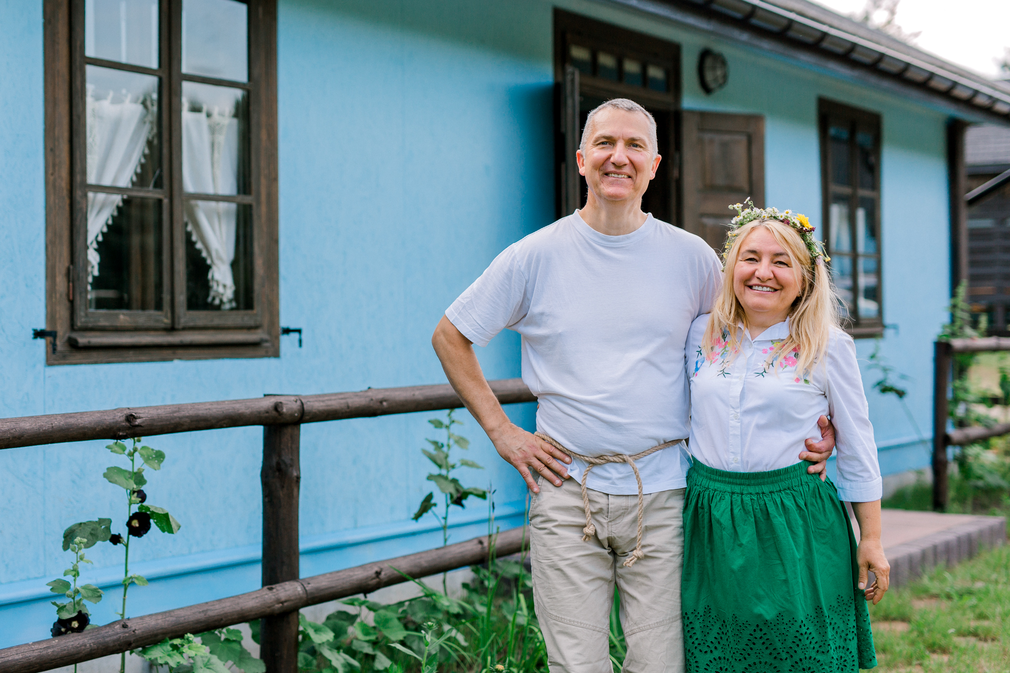 Para w tradycyjnych strojach przed niebieskim wiejskim domem, kobieta w wianku z kwiatów, idealne na zdjęcia z wakacji agroturystycznych.