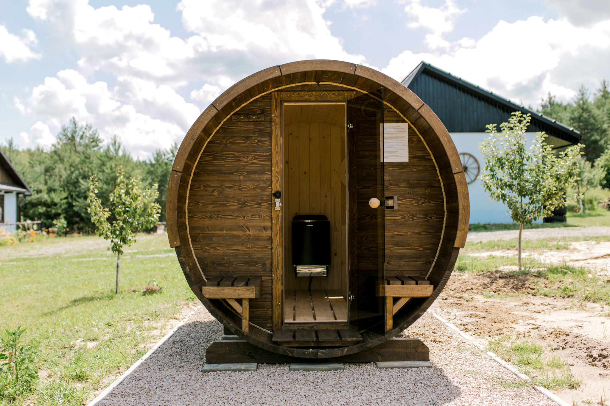 Zewnętrzny widok sauny beczkowej z otwartymi drzwiami, na zielonej trawie otoczonej lasem, z nowoczesnym piecem wewnątrz dla komfortowego saunowania na świeżym powietrzu.