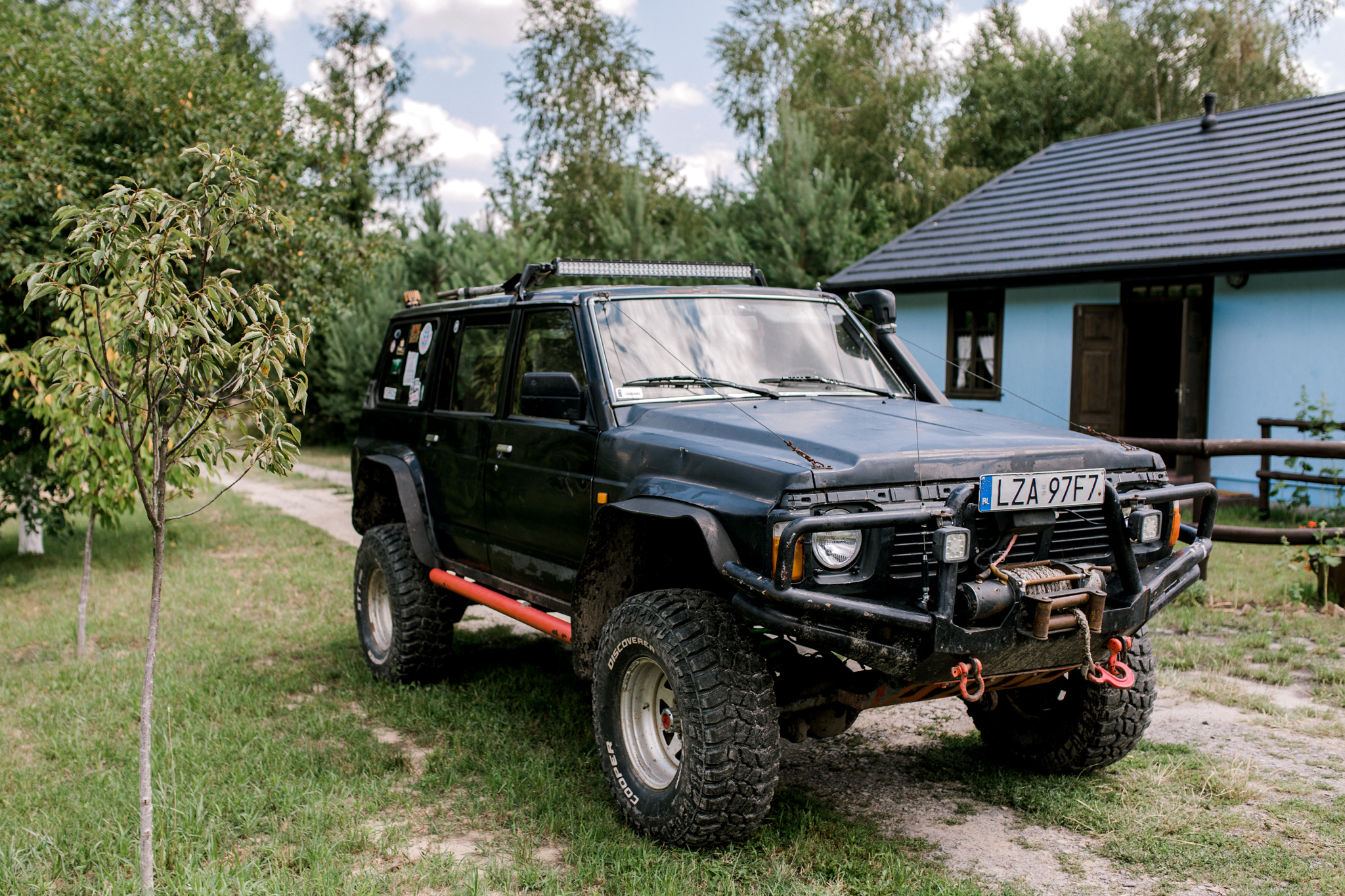Czarny Nissan Patrol off-road z wyciągarką i dużymi oponami terenowymi zaparkowany przed wiejskim domem w zielonym otoczeniu lasu i ścieżki.