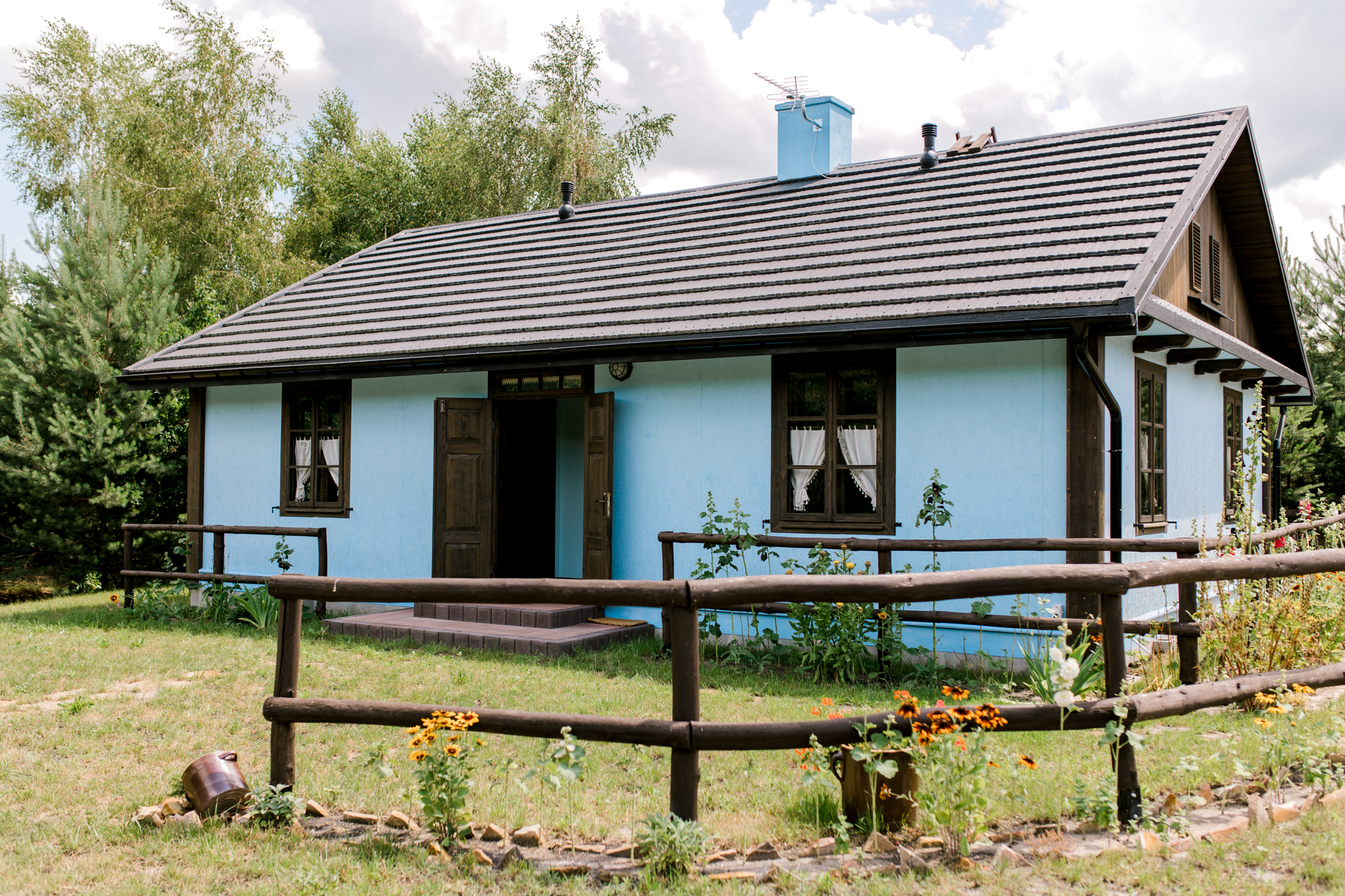 Niebieski domek letniskowy na wsi z kwiatami przy drewnianym płocie, otwartymi drzwiami i zielonym ogrodem na świeżym powietrzu