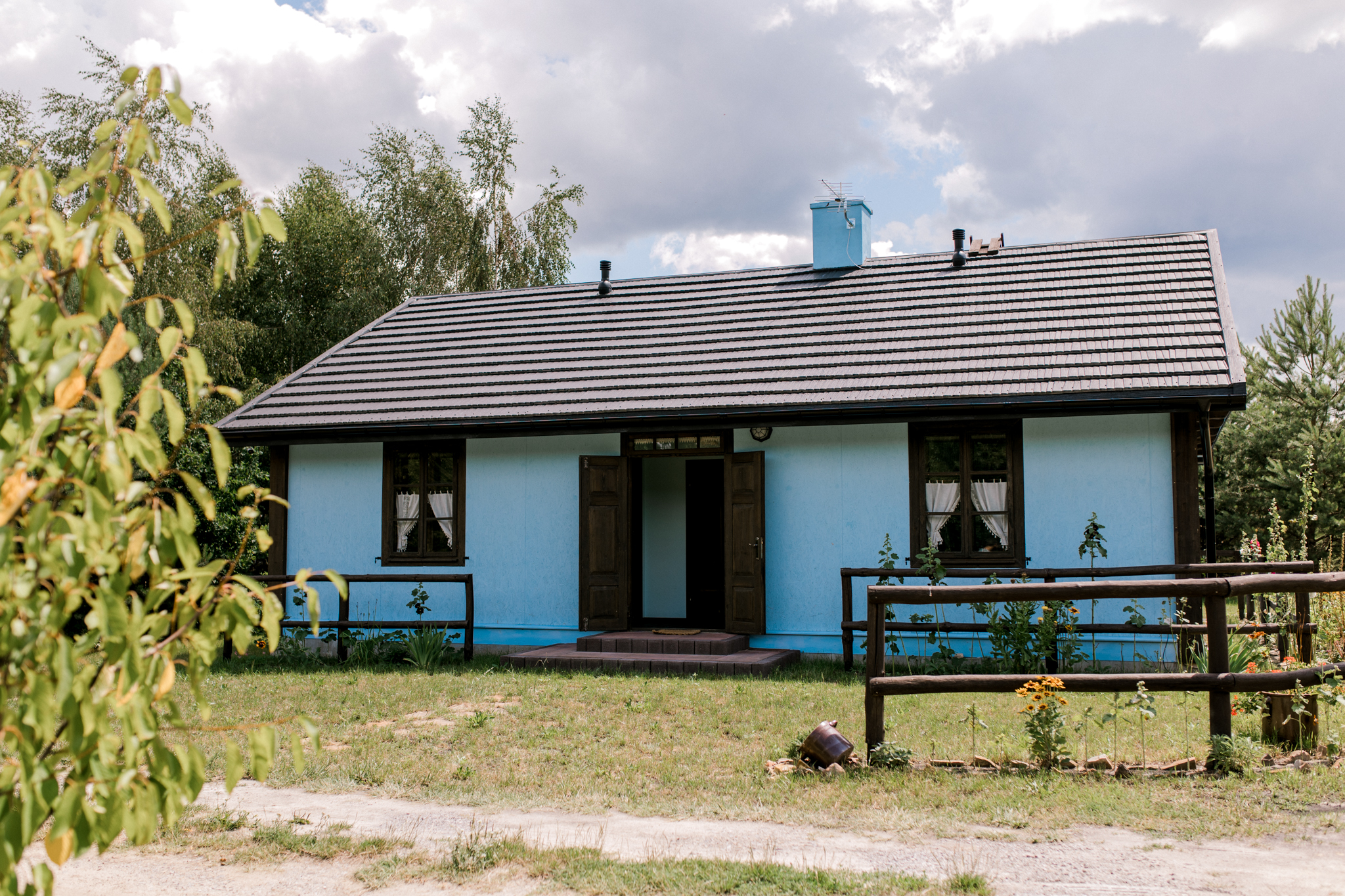 Rustykalna starodawna chata w kolorze niebieskim z drewnianym płotem, roślinami i widokiem na zielony ogród ruralny