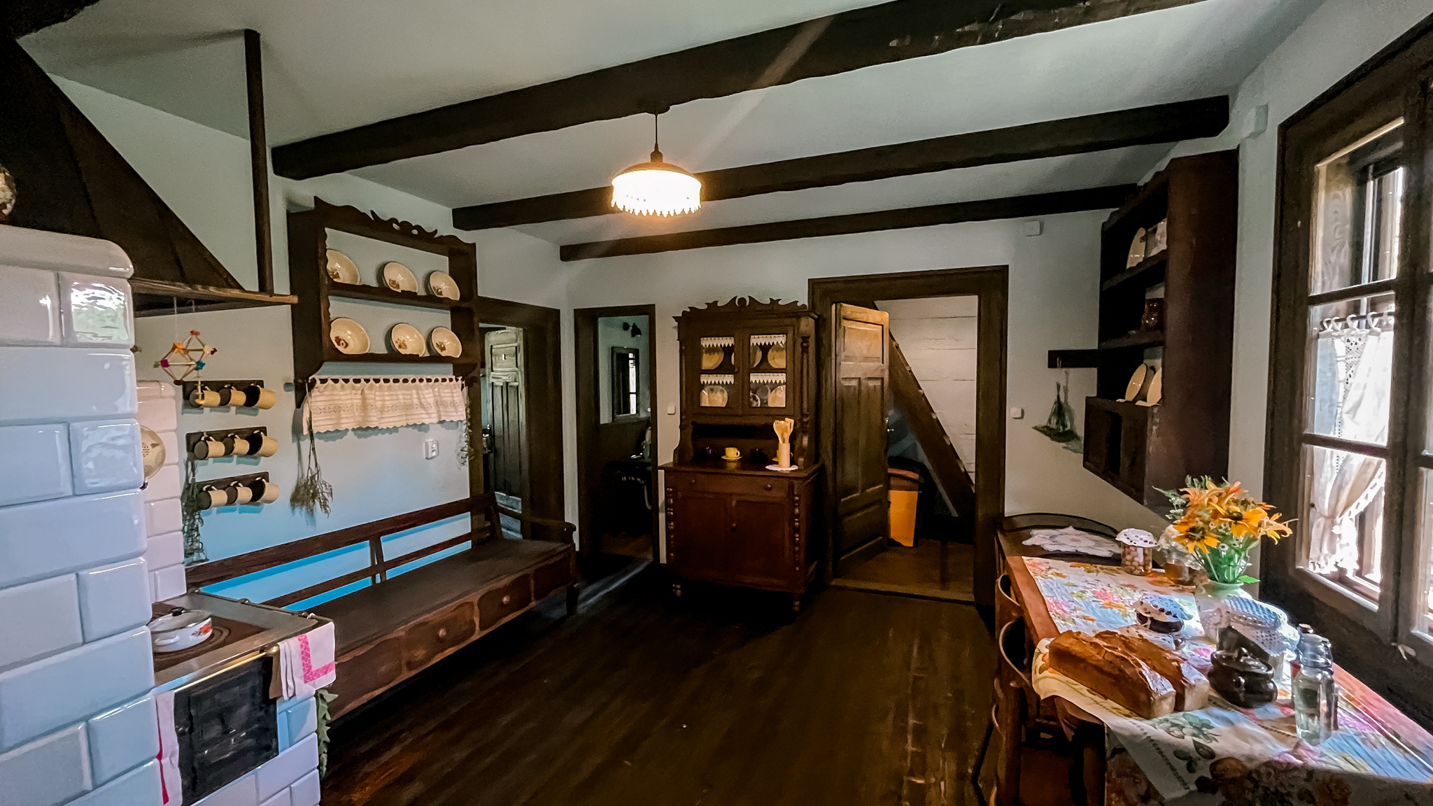 Wiejska kuchnia z drewnianym stołem, regałami z naczyniami, bukietem kwiatów i schodami w tle pod belkowanym sufitem.