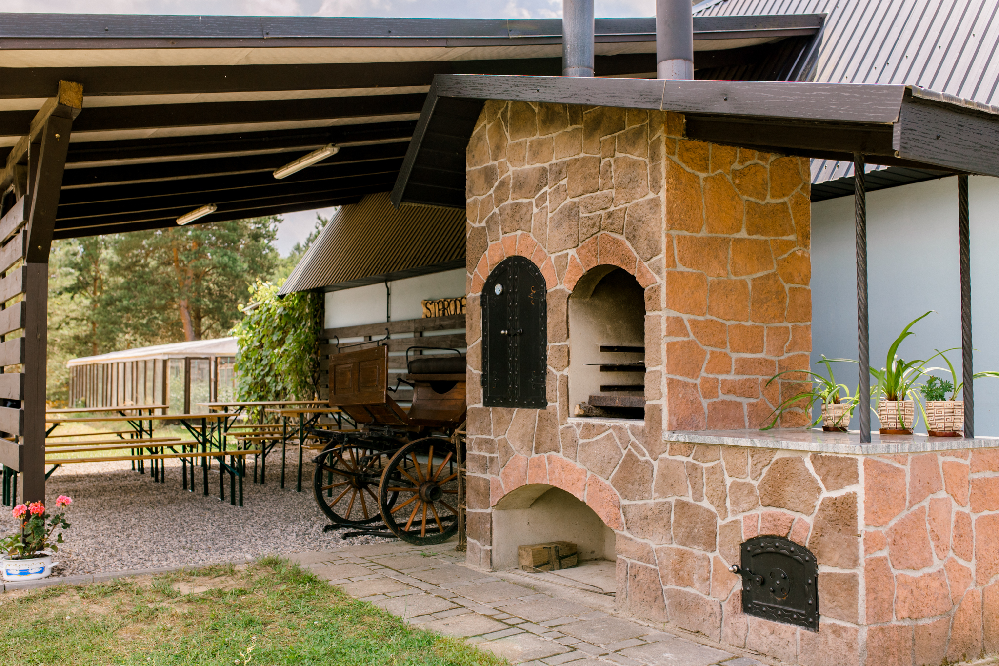 Kamienny piec chlebowy z dwoma otworami pod zadaszeniem, obok tradycyjny wóz z drewnianymi kołami, stoły piknikowe i zielone rośliny w tle.